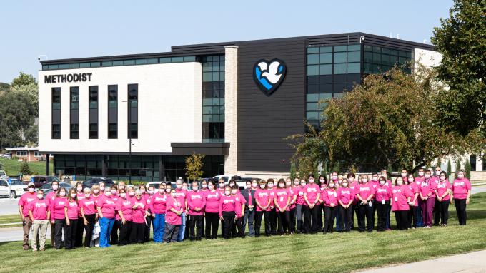 A large group of people wearing pink shirts stand in a line on the lawn of the Methodist Jennie Edmundson Hospital. 