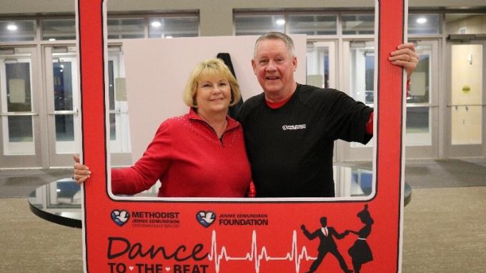 A woman in a red shirt and a man in a black shirt are standing behind a red photo prop that looks like a photo frame. The frame says “Dance to the Beat” on it. 