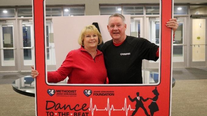 A woman in a red shirt and a man in a black shirt are standing behind a red photo prop that looks like a photo frame. The frame says “Dance to the Beat” on it.