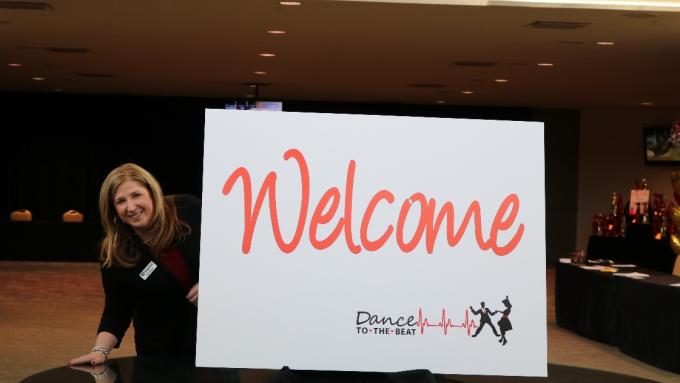 A woman stands next to a sign that reads “Welcome: Dance to the Beat.” 